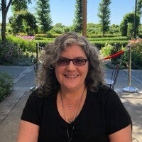 White female with black/gray curly hair, wearing black v-neck top, sitting in front of grassy area with flowers