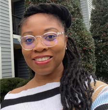 Portrait of Abisola Osinuga. Dr. Osinuga is a Black woman with long black dreadlocks tied back. She is wearing wire rimmed glasses and a black, tan and white sweater and is smiling at the camera.