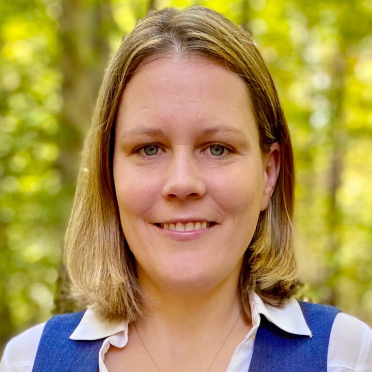 Portrait of Jaimee Zeyzus. Jaimee is a white woman with straight shoulder-length blond and brown hair. She is wearing a white collared shirt and blue vest and smiling at the camera in a grove of trees.