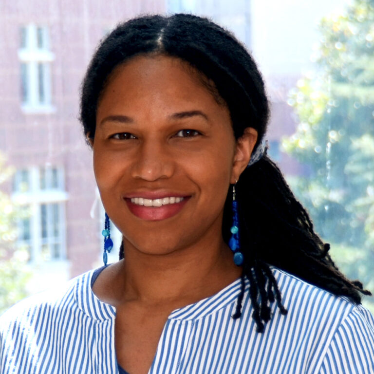 Portrait of Tara Carr. Tara is a Black woman with long twists, tied back in a ponytail. She is wearing blue dangly earrings and a blue striped blouse and smiling at the camera in front of a window.