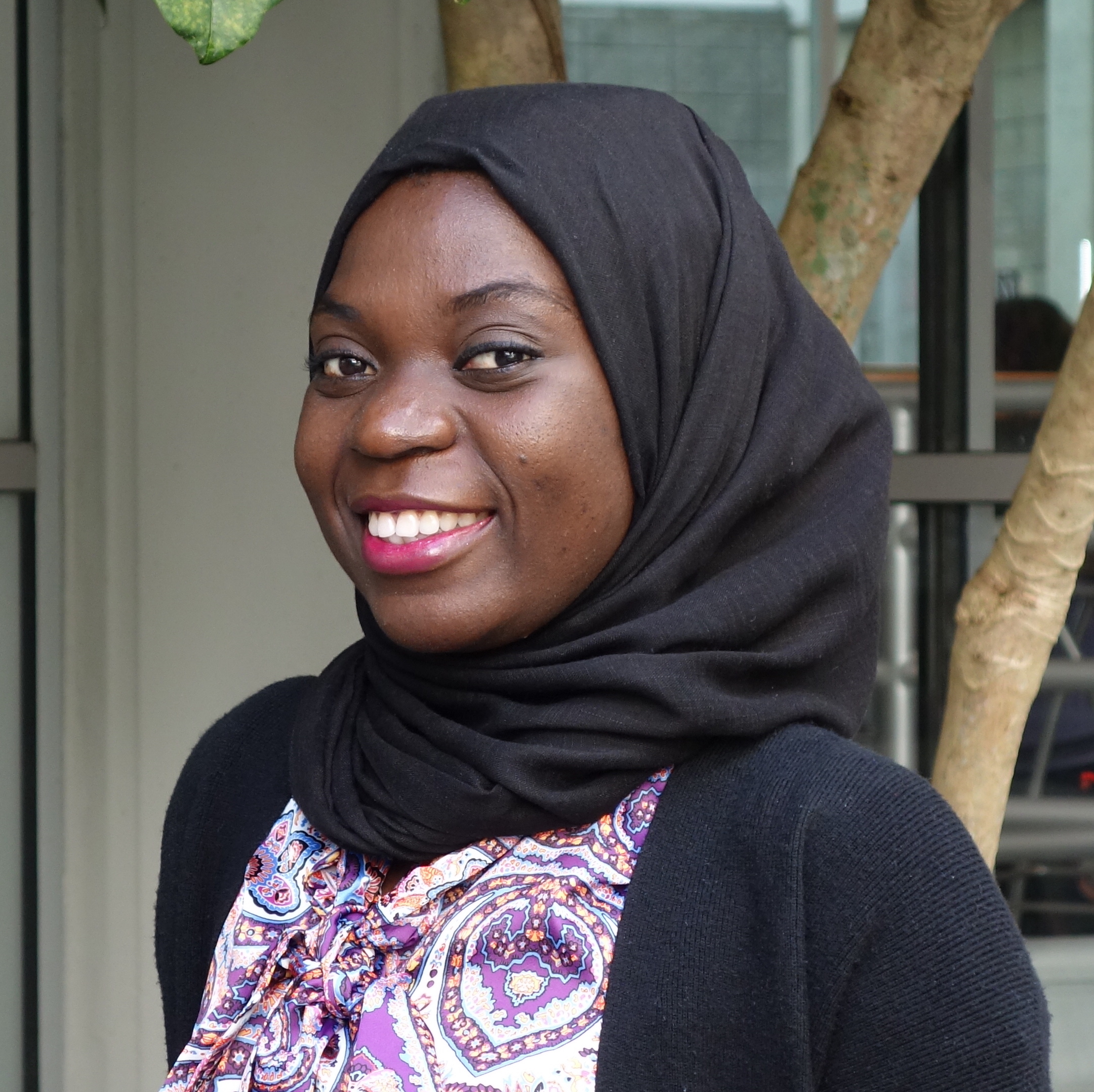 Portrait of Bola Yusef. Bola is a Black woman wearing a black headscarf and purple patterned shirt. She is standing in front of a tree and smiling at the camera.