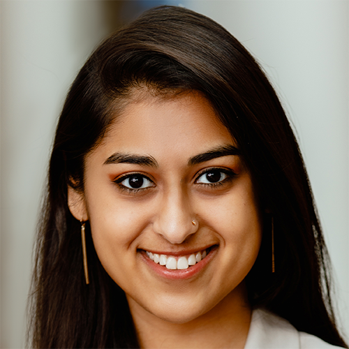 Portrait of Abhigna Rao. Abhigna is a woman with straight brown hair. She is wearing a white shirt and is smiling at the camera. She is posed against a white/gray background.