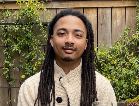 Portrait of Bernard Coles IV. Bernard is a male with long dark hair that extends past his shoulders. He is wearing an off white sweater with dark buttons and is smiling at the camera.