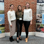 Three women, members of the Abacus Evaluation team, stand together between two standing banners for the Carolina Across 100 Our State, Our Wellbeing program.