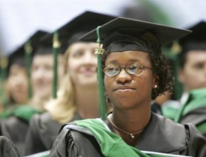 A close up of a smiling female graduate wearing a black cap and gown and green sash. Other graduates are behind her. Online Course offerings