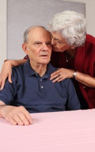 An elderly man looking confused and seated at a table. An elderly woman is leaning over to kiss him on the cheek.
