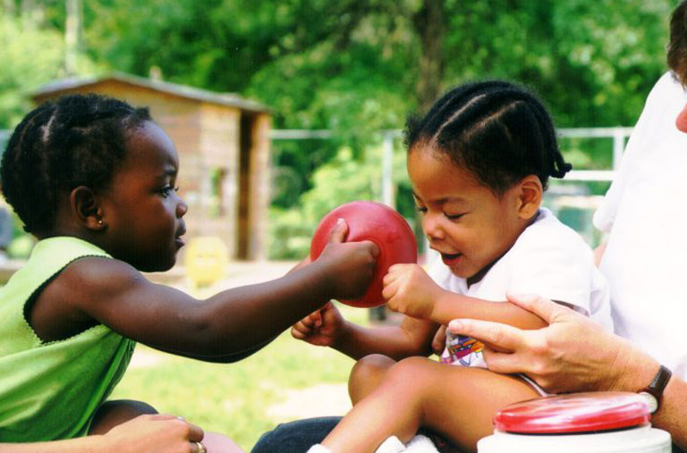 Two preschool-aged children passing a red ball. One is on an adult's lap and the adult is helping her catch the ball