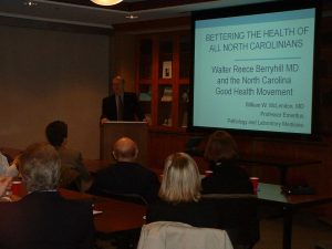 Dr. McLendon at a podium with a projector screen displaying the title of his talk, "Bettering the Health of all North Carolinians." A few audience members are in the foreground.