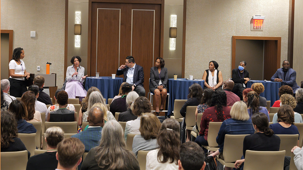 A row of panelists sit at a cloth-draped table. Multiple rows of seated audience members are facing the panelists.