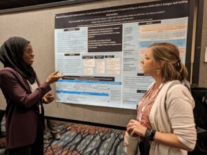 A Black woman wearing hijab explains a research poster to a white woman in a hoodie.