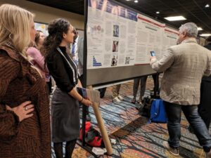 Two white woman stand beside a research poster as a white man takes a photo of the poster.