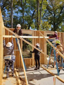 Five volunteers in hard hats work together to raise a large beam in a house being built.
