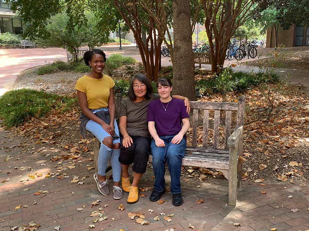Dr. Haiyan Fu sitting on a wooden bench with two lab members on UNC campus