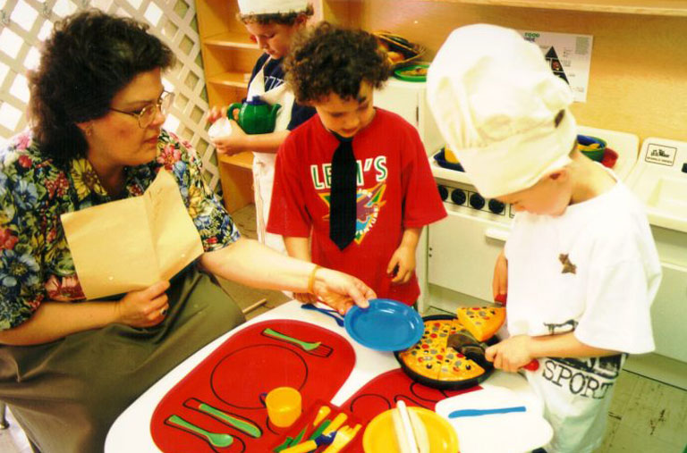 Teacher and two preschool-aged boys in a kitchen center in a preschool classroom. One boy is dressed as a chef with a white hat and is cutting a pretend pizza while the adult holds a plate