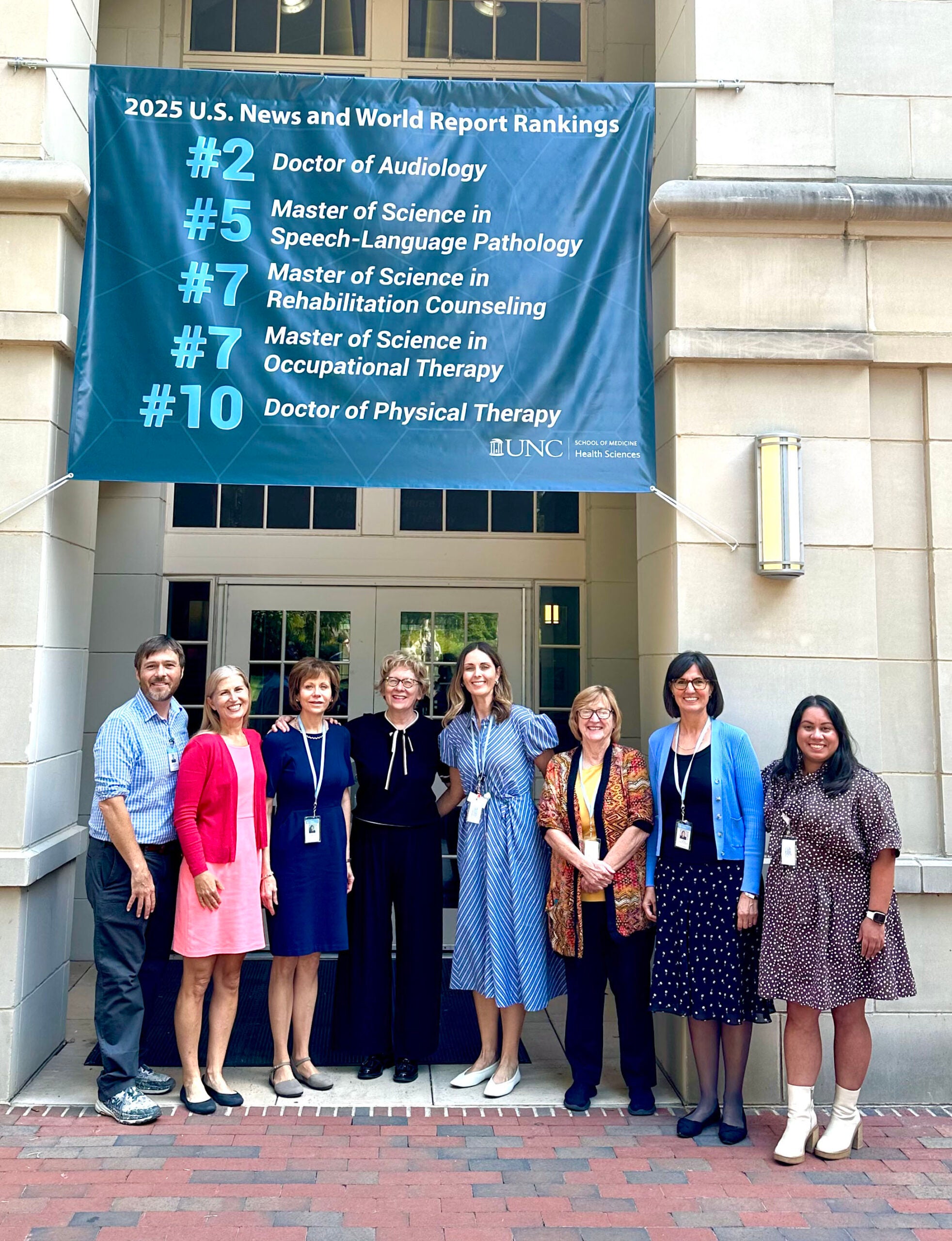 CRMHC Faculty Standing outside the front of Bondurant Hall under a banner with US News and World Report Rankings.