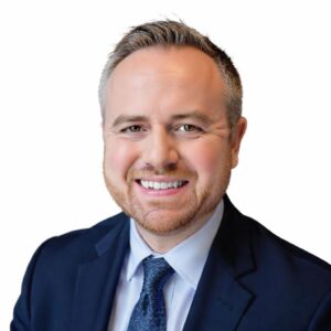Kyle Cooper headshot, white man with short light brown hair wearing a dark blue suit with a light blue shirt and dark blue tie