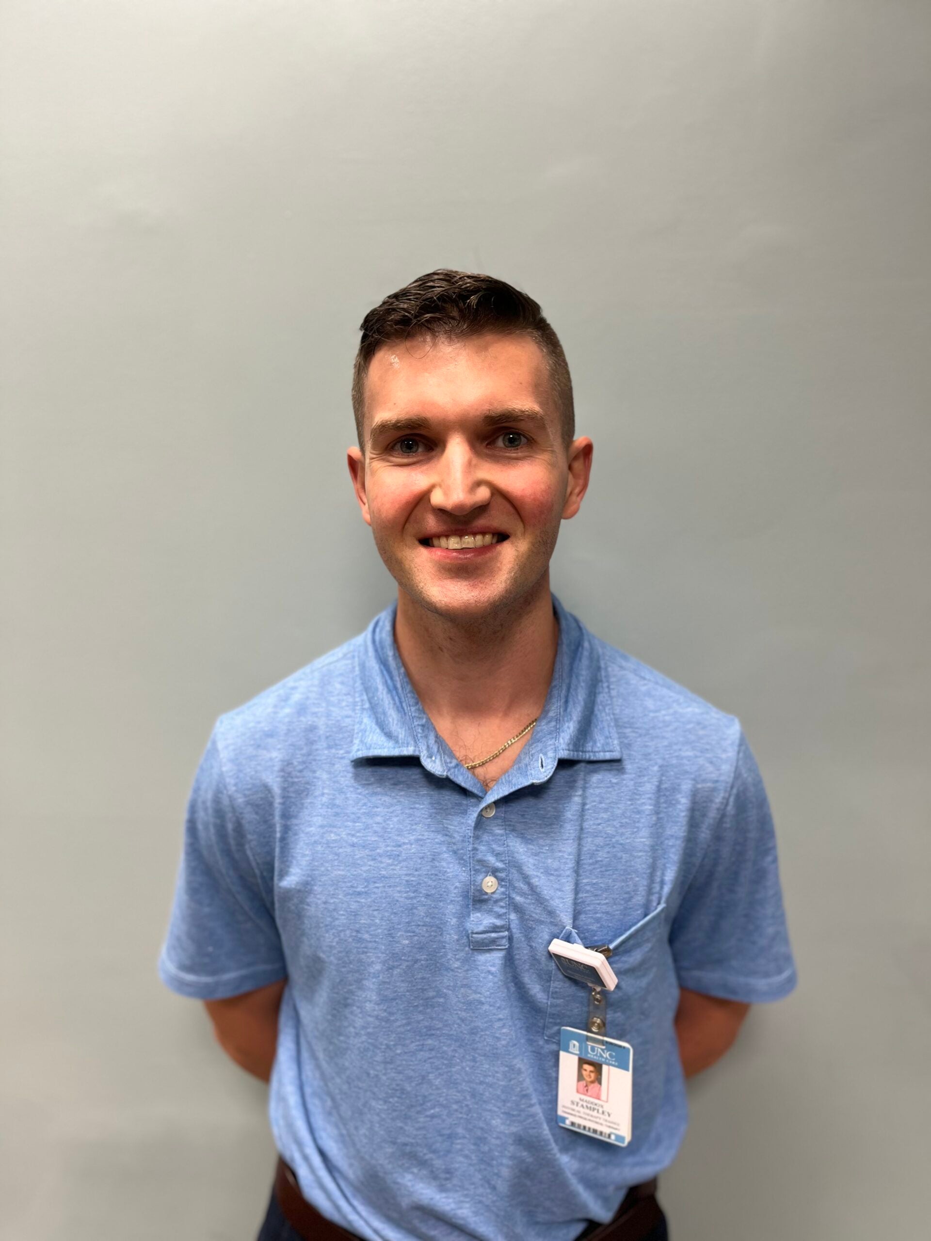 Maddox Stampley in a blue collared shirt, smiling in front of a grey background.