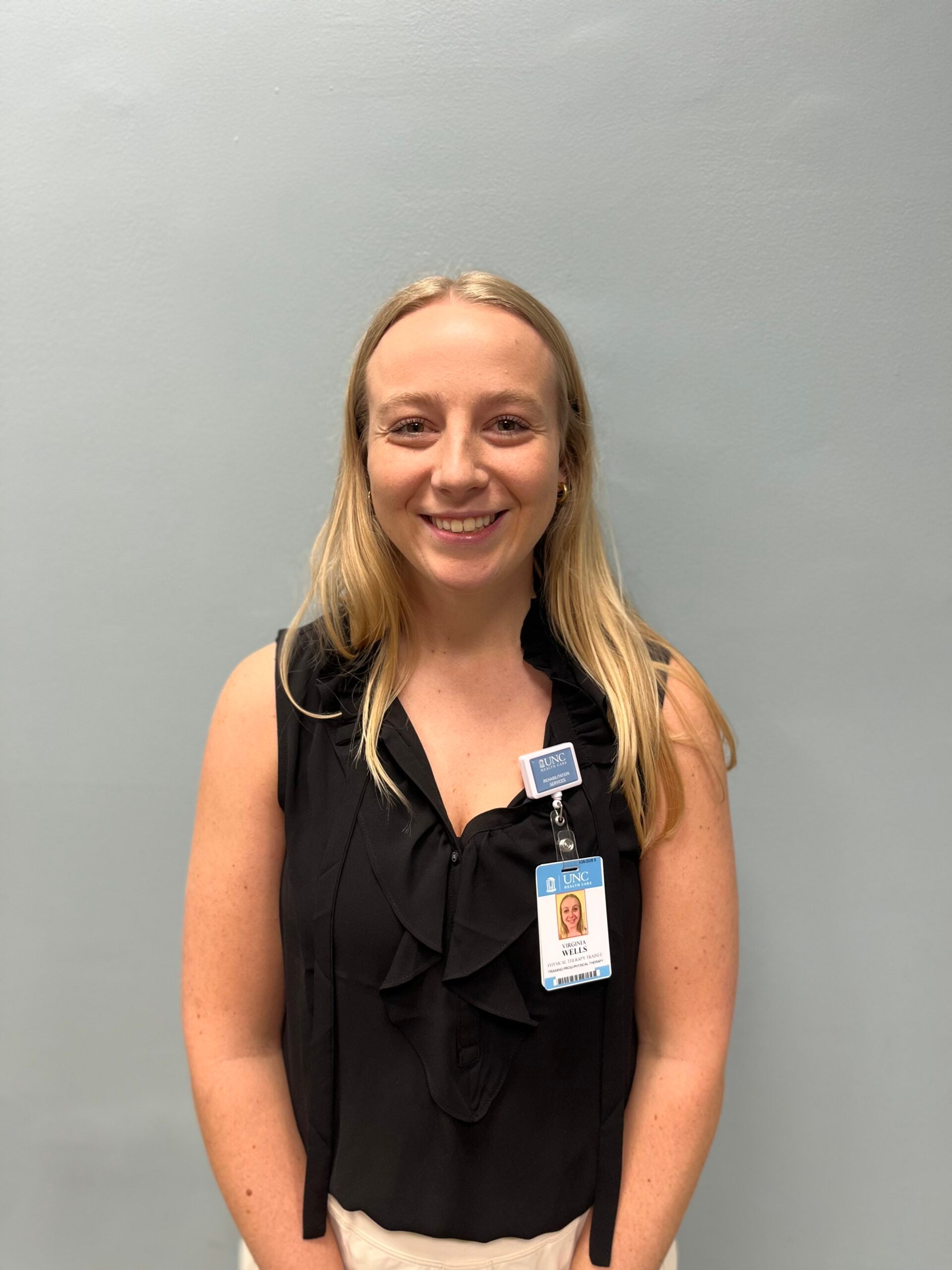 Virginia Wells in a black top smiling in front of a grey wall.