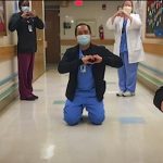 A team of 5 UNC Health employees, all wearing scrubs and disposable masks, stand or kneel feet apart from one another while forming heart symbols with their hands.