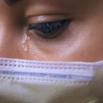 A healthcare worker wearing a disposable mask looks down. A tear or drop of sweat falls the corner of her eye.