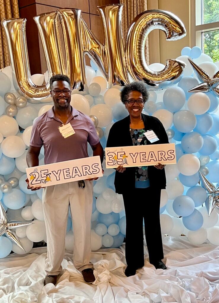 Rodney Gilmore and Marguerite Applin standing in front of a wall of balloons and holding signs noting the length of their service at UNC