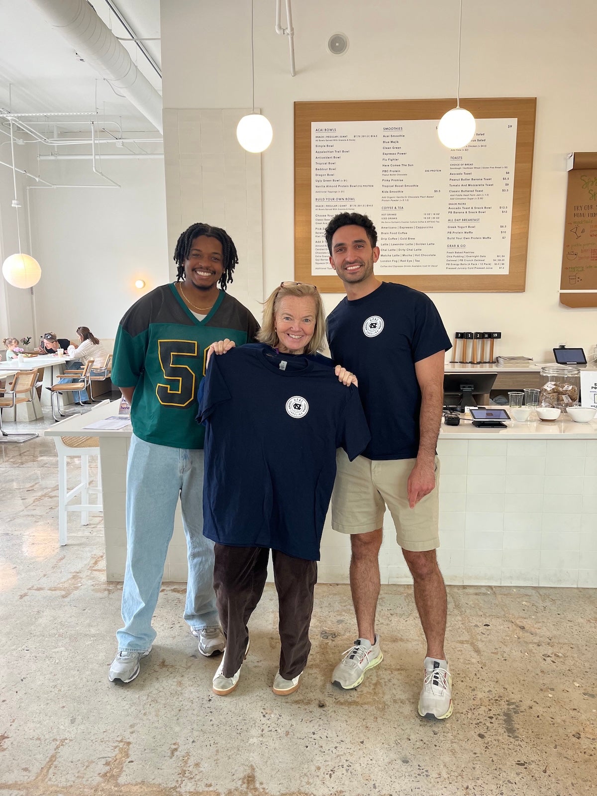 A group photo of three people. The woman in the middle is holding up a t-shirt.