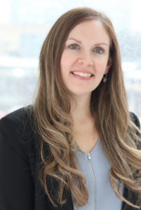 Studio headshot of a healthcare professional wearing a black blazer and light blue top against a softly blurred indoor background.