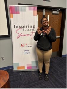 A woman pictured supporting her sorority with a hand gesture, in front of a banner that reads "Inspiring Futures Summit"