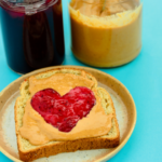 A slice of bread with peanut butter and Jelly on top shaped like a heart with jars of PB and Jelly placed behind it