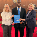 Three individuals stand in a courtroom holding a “Building a World Without NEC” sign beneath the North Carolina state seal.