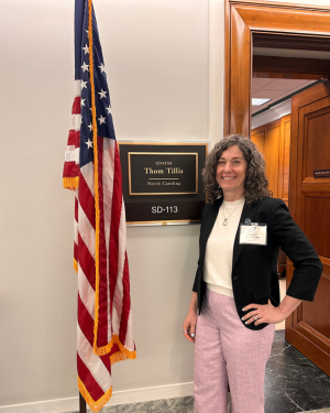 Dr. Maureen Petersen stands in a hallway beside a U.S. flag and a wall plaque reading “Thom Tillis, North Carolina, SD-113,” with a wood-paneled office doorway behind.