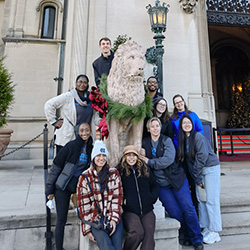 Pharmacology 2nd year students group photo at the Biltmore Estate, Asheville NC. There are seasonal Christmas wreaths decorationing a stature of a lion the group stands around and a fancy light pole.