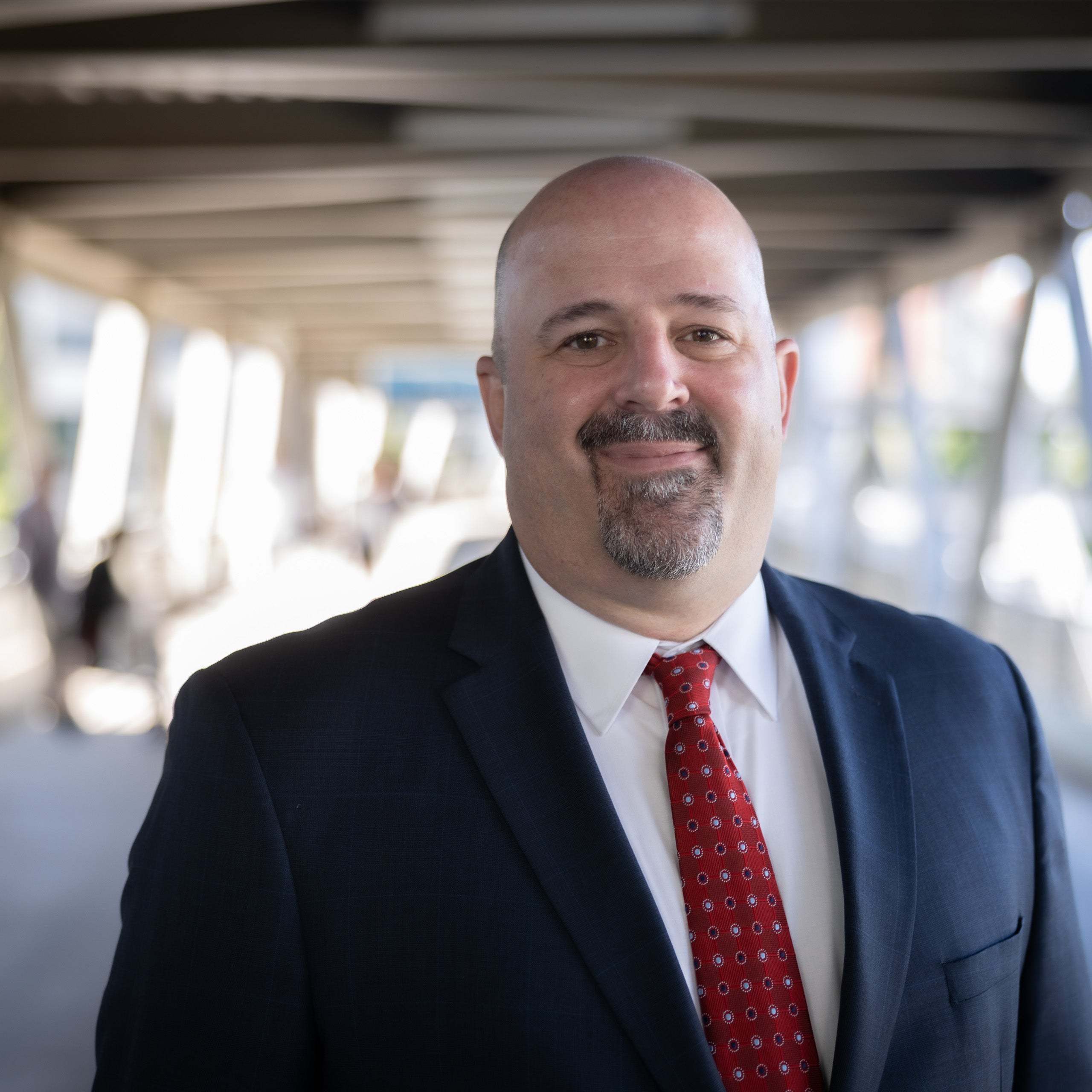 Dan Boughton headshot. Bald male with brown eyes and goatee and mustache. He is wearing a white shirt, red tie and black jacket.