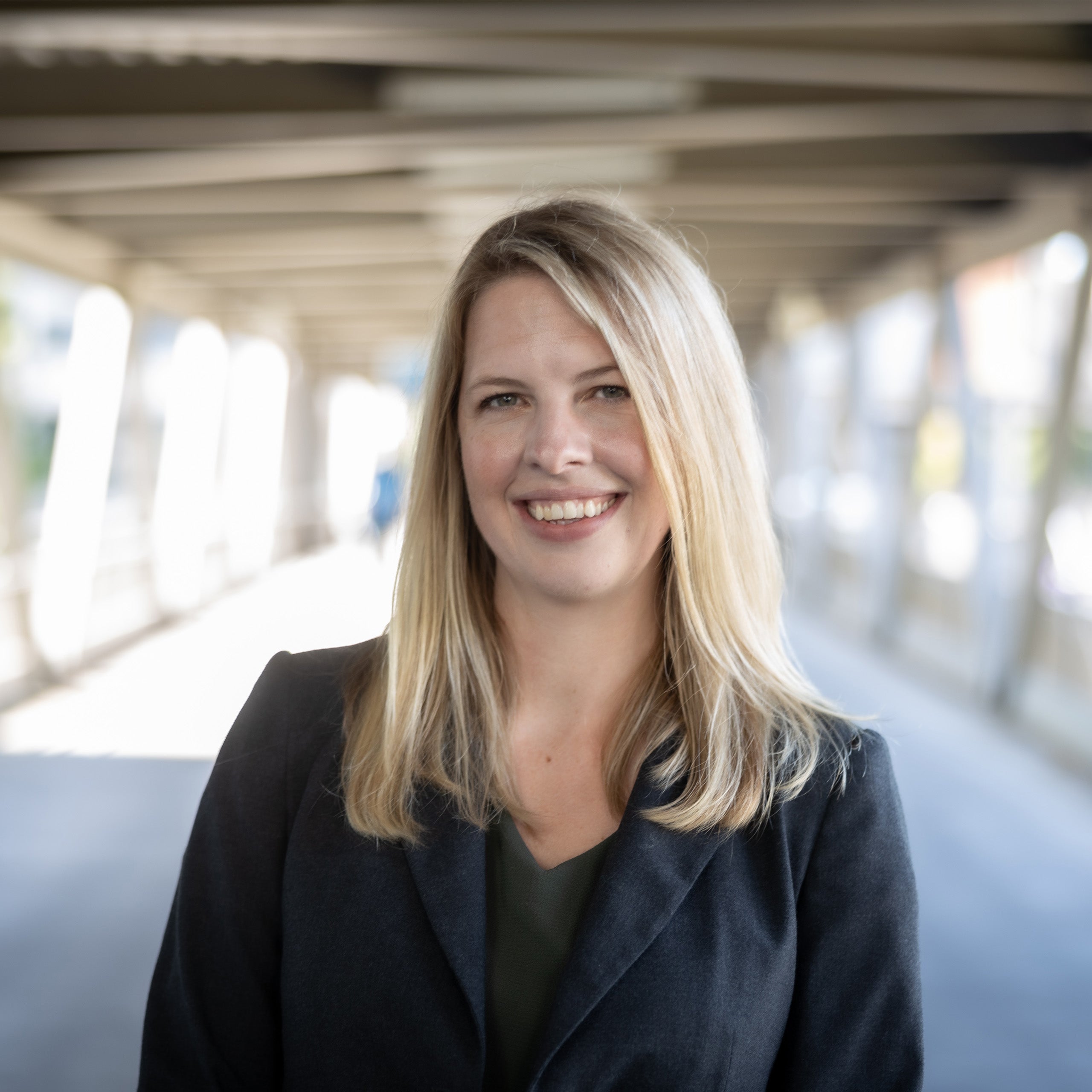 Kristen Olinger headshot. Female with long blond hair and light eyes smiling at the camera. She is wearing a black blouse and a suit jacket.