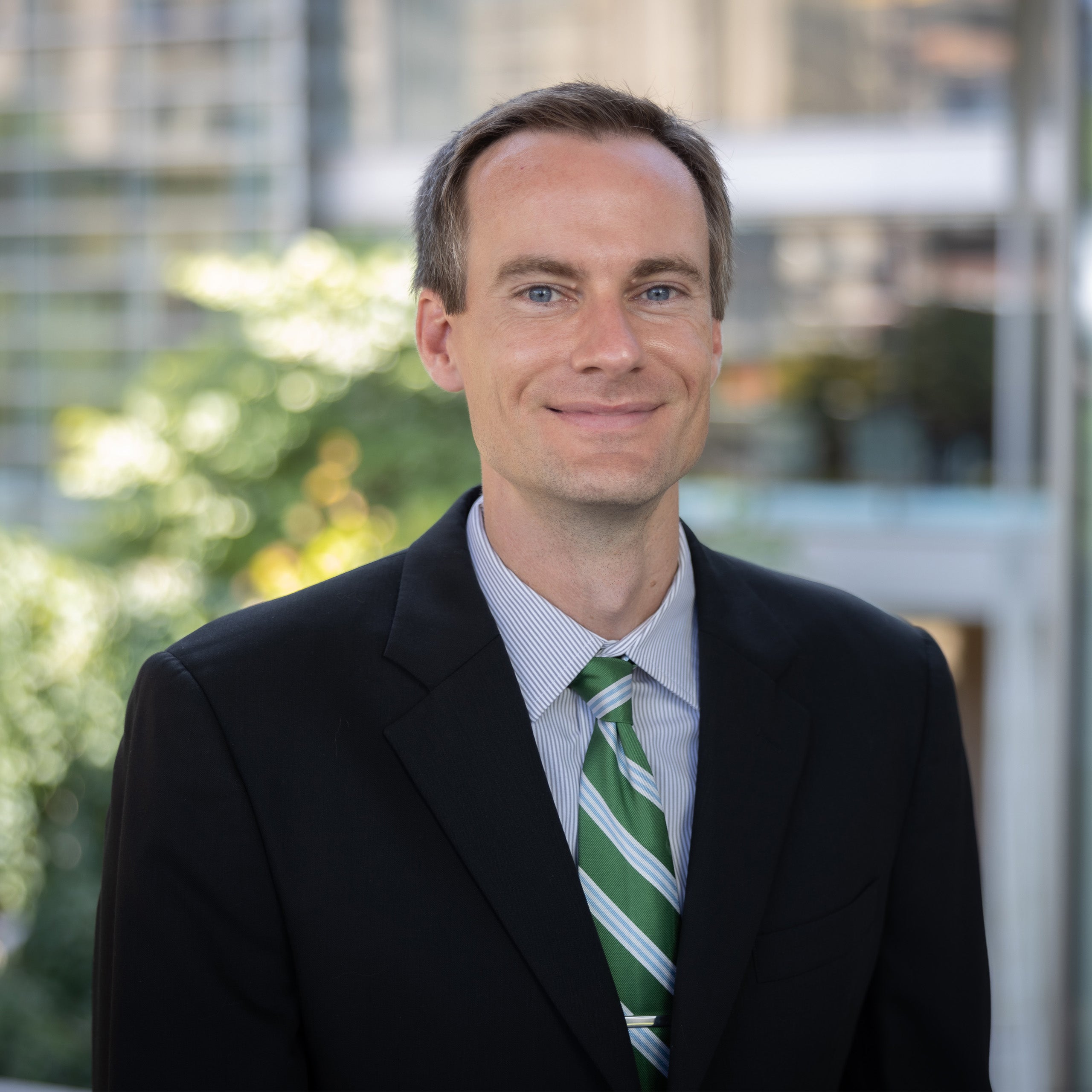 Chris young headshot. A male with short brown hair and blue eyes is smiling at the camera. He is wearing a white with a blue strip shirt, a green tie, and a black suit jacket.