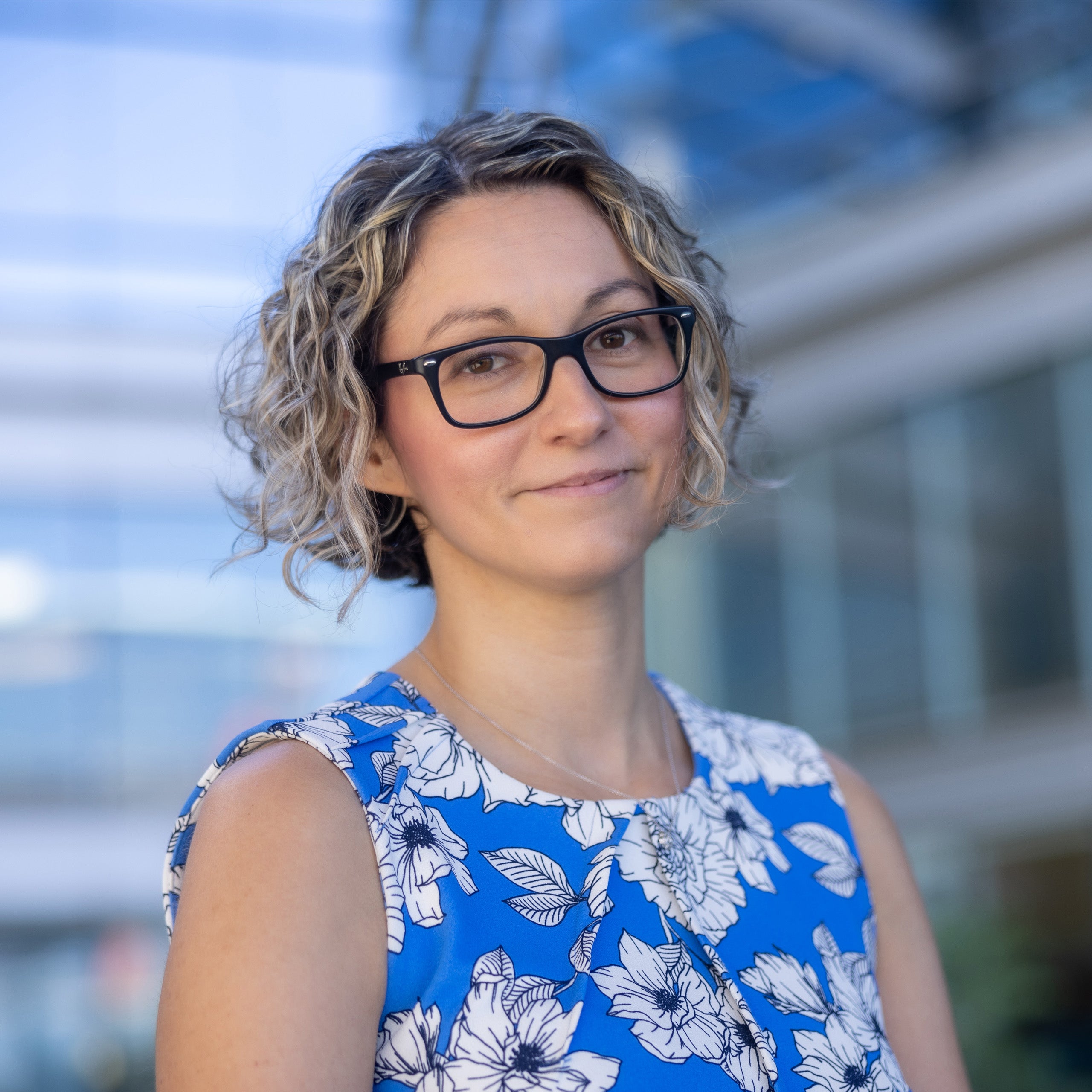 Genevieve Woodard, MD, headshot. Female short curly brown hair with blonde highlights, brown eyes, and wearing glasses, smiling at the camera. She is wearing a sleeveless blue dress with white flowers.