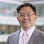 Yueh Lee, MD, PhD, headshot. An Asian male with short black hair and brown eyes, wearing glasses, is looking at the camera. He is wearing a white shirt, a pink tie, and a grey suit jacket.