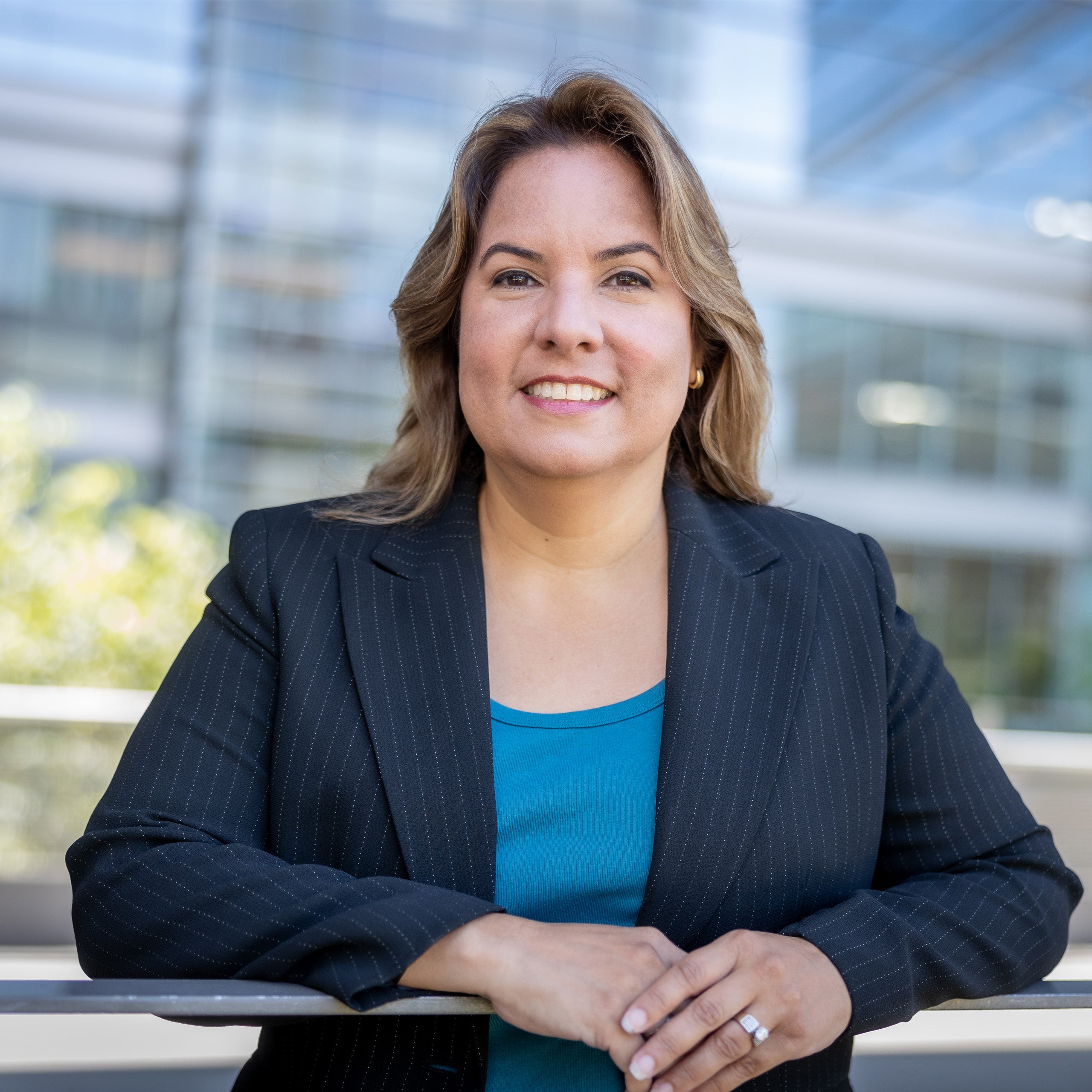 A female with honeybrown hair to her shoulders and dark brown eyes is smiling at the camera. She is wearing a dark striped suit and blue shirt with her hands clasped in front of her on a rail. She is standing against a glass building.