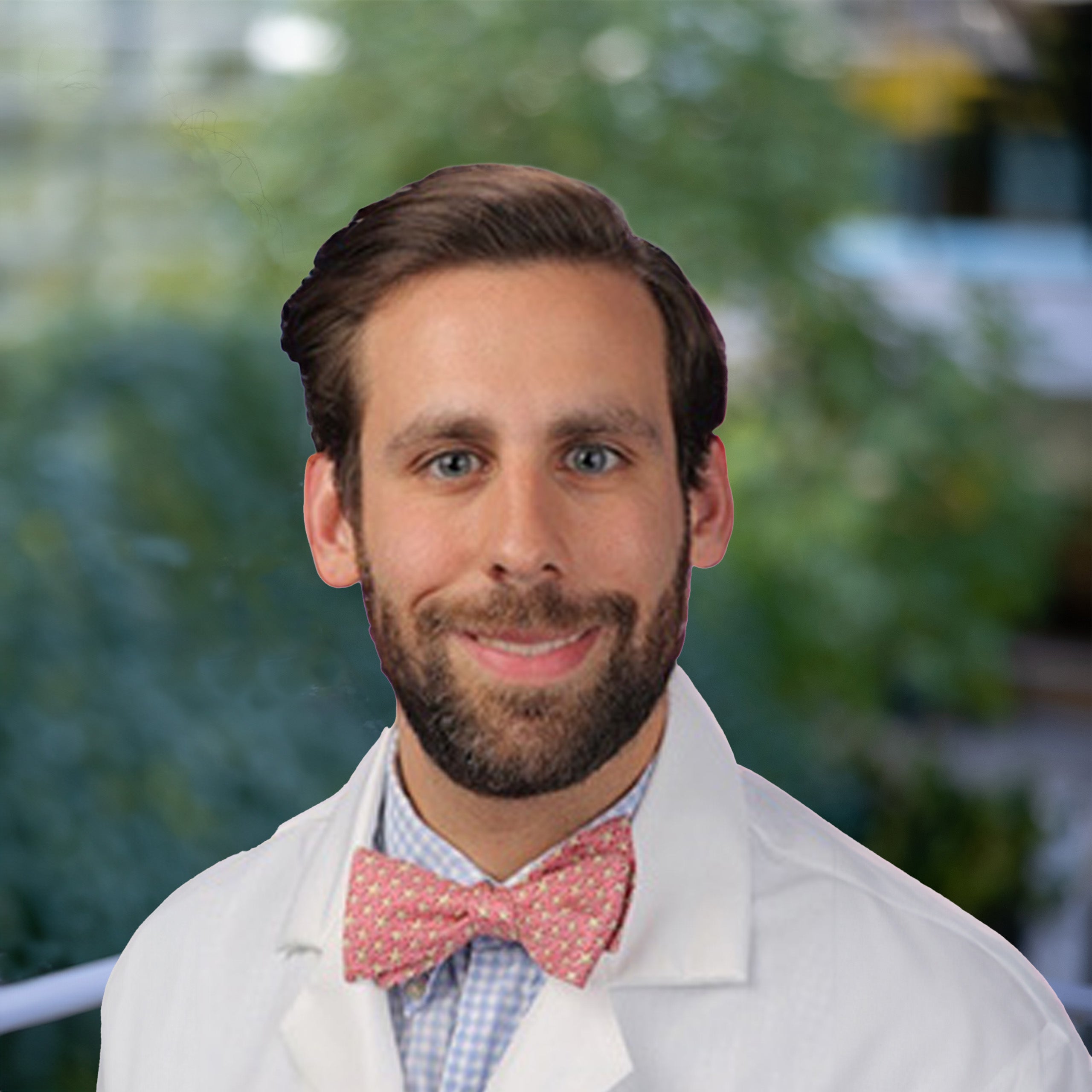Will Pryor's headshot. A male with light brown hair, a beard, and blue eyes is smiling at the camera. He is wearing a blue and white checkered shirt with a pink bow tie and a white doctor's coat.