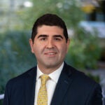 Nima Kokabi's headshot. A male with dark brown hair and eyes smiling at the camera. He is wearing a white shirt, bright yellow tie, and dark blue suit jacket.
