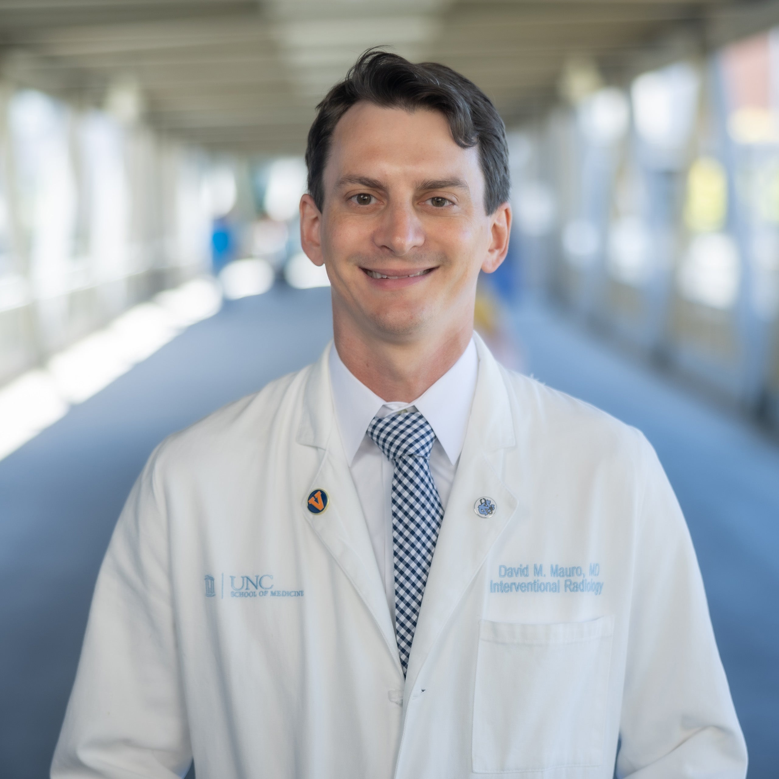 David Mauro headshot. Male with brown hair and eyes is smiling at the camera. He is wearing a white shirt, a black checkered tie, and a white doctor's coat.