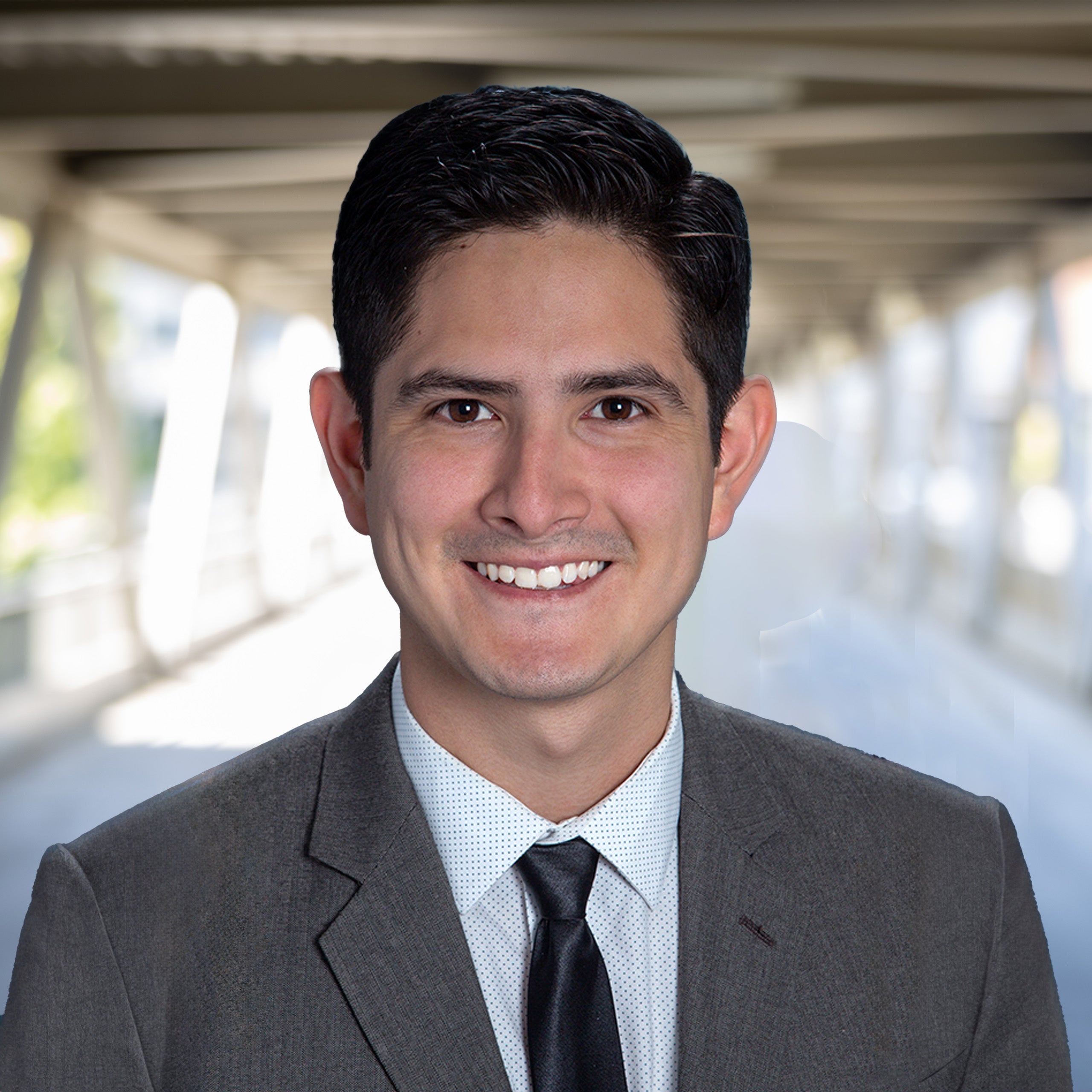 Alex Villalobos's headshot. A Male with short dark brown hair and eyes smiling at the camera. He is wearing a white shirt, black tie and grey suit jacket.