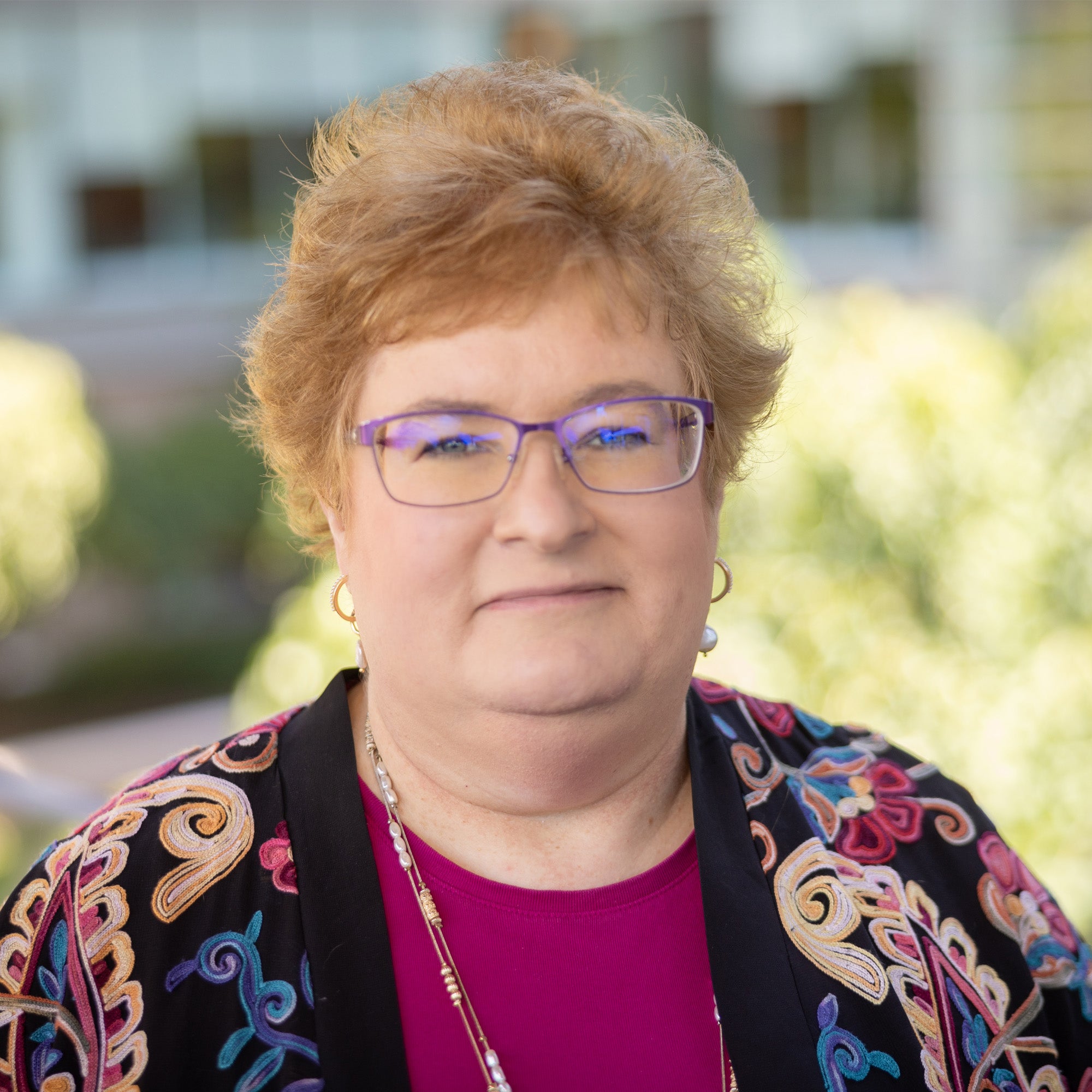 Michele Clark headshot. White female with short red hair and green eyes with glasses. She is wearing a pink shirt with a colorful cardigan.