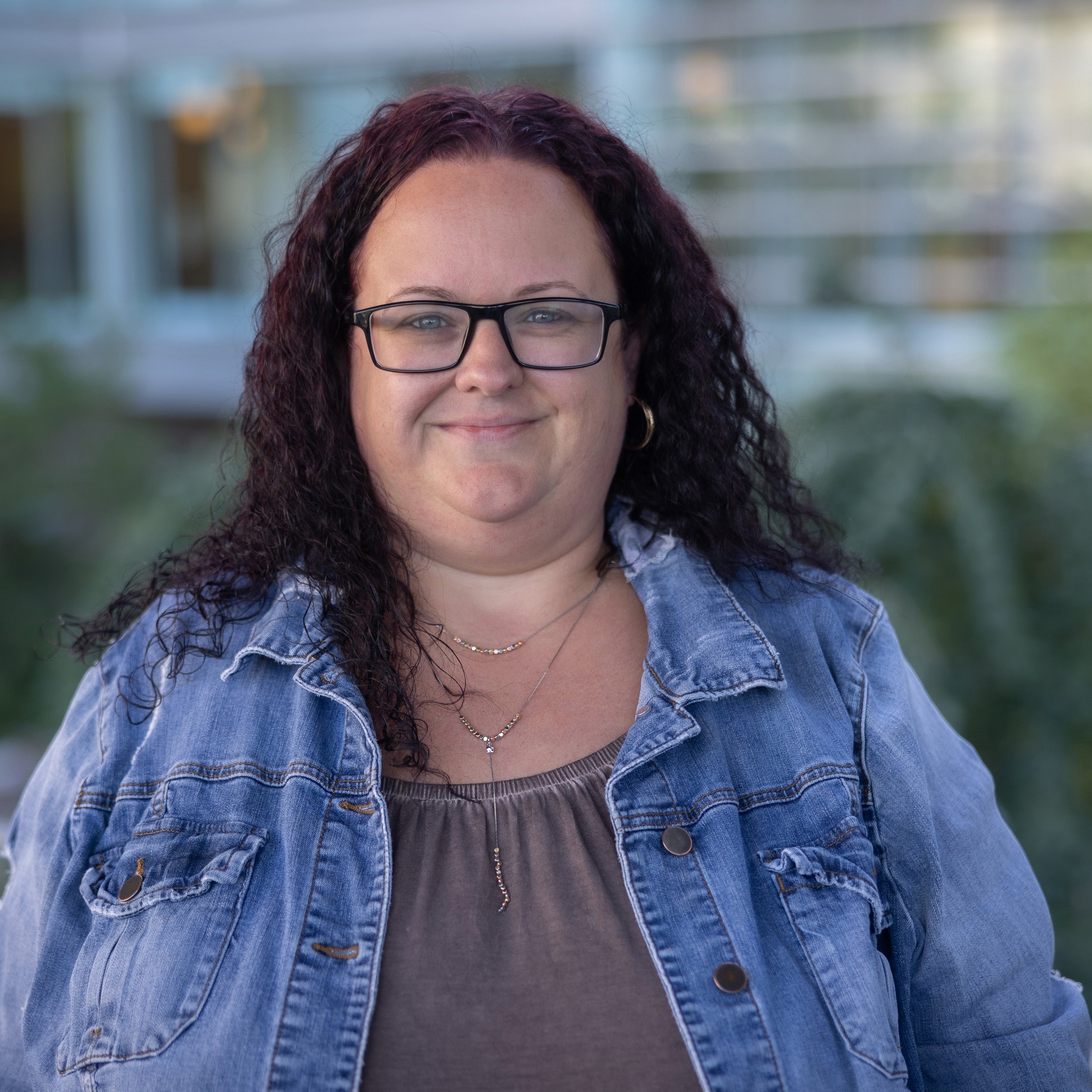 Sarah Edwards headshot, Long brown curly hair, and green eyes with glasses, smiling at the camera. She is wearing a grey blouse with a jean jacket.