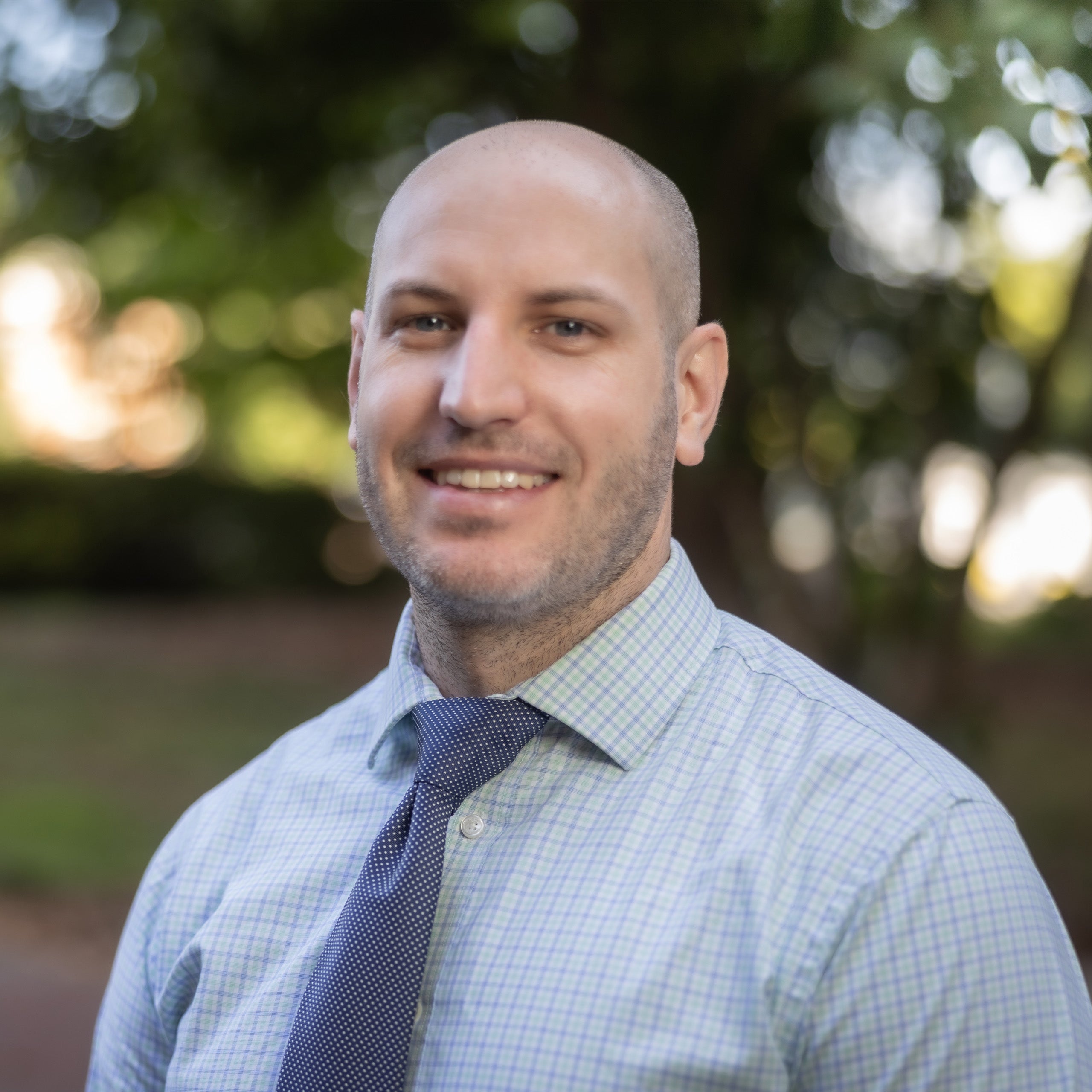 David Bartlett, MD, headshot. A male with a shaved head and light eyes smiling at the camera. He is wearing a checkered shirt and blue tie.