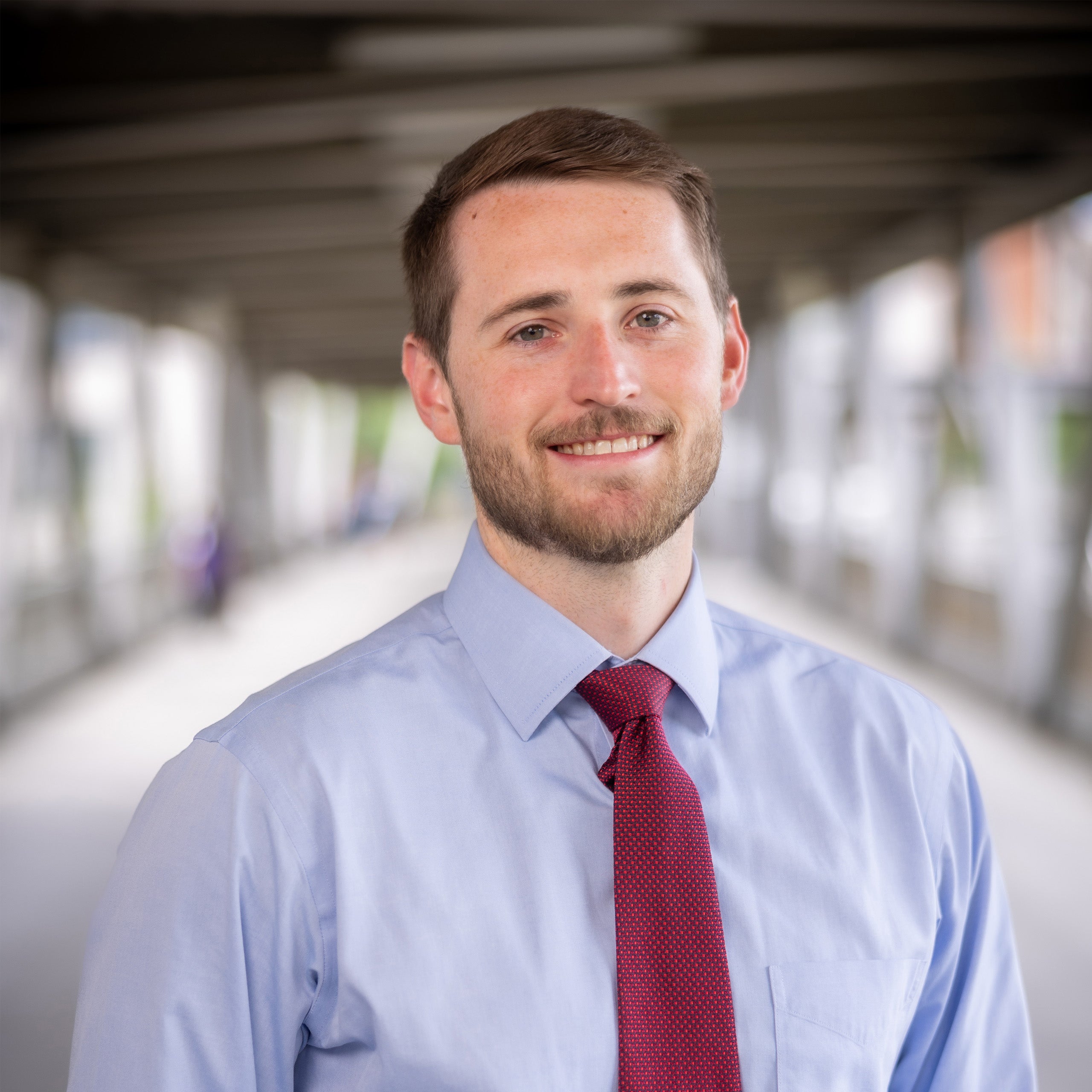 Timothy brown headshot. Male with brown hair and a beard with hazel eyes smiling at the camera. He is wearing a blue shirt and red tie.
