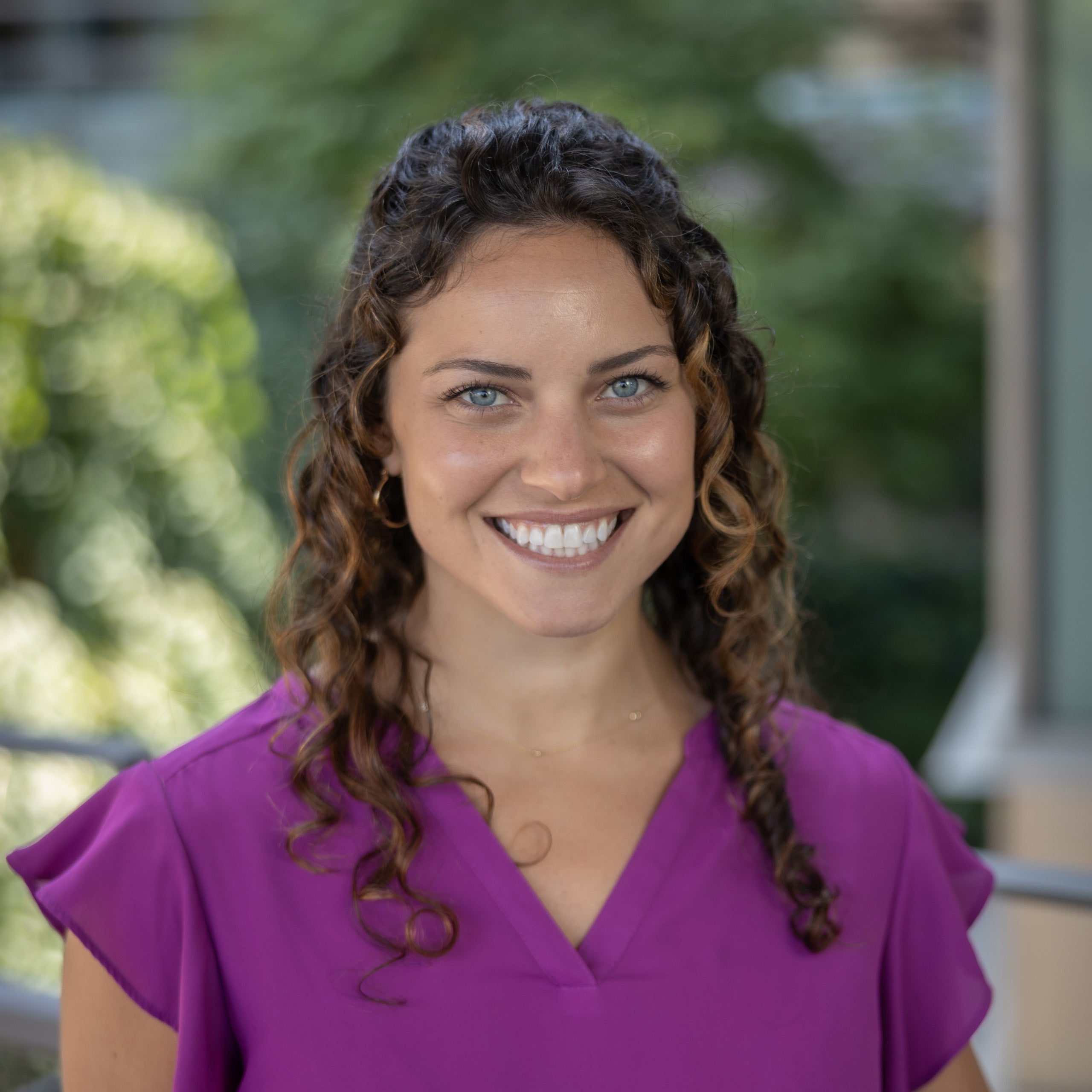 Cristina Guzzetti headshot. A female with curly brown hair and blue eyes is smiling at the camera. She is wearing a purple blouse with flouncy sleeves.