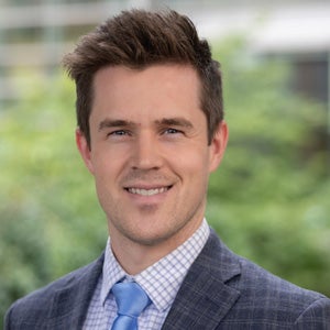 Matthew Maclean's headshot. Male with short, spiky brown hair and light eyes. He is wearing a checkered suit, shirt, and a light blue tie. He is standing against an outdoor backdrop.