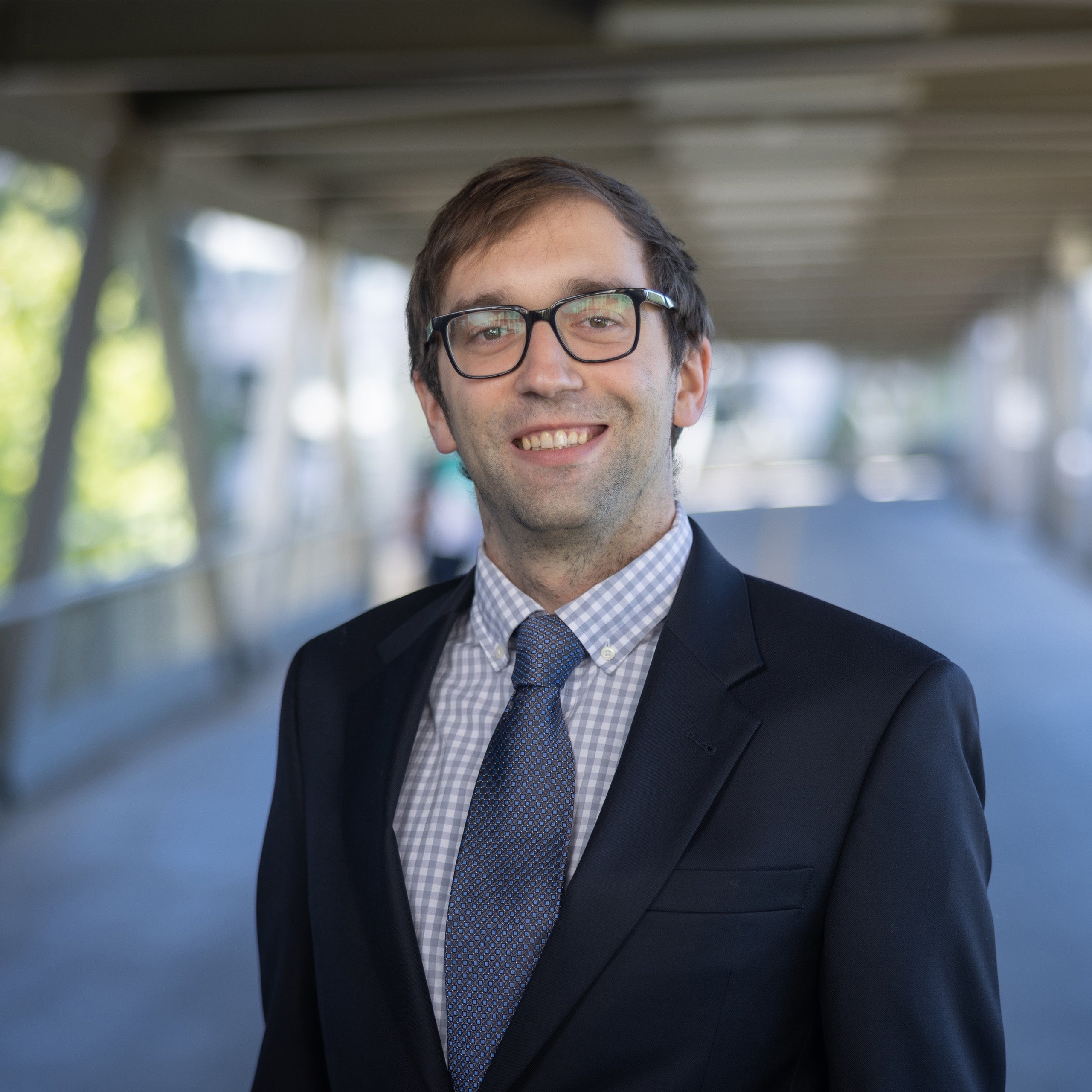 David Sailer headshot. Male with short brown hair and eyes wearing glasses smiling at the camera. He is wearing a white and blue checkered shirt, blue tie and black jacket.