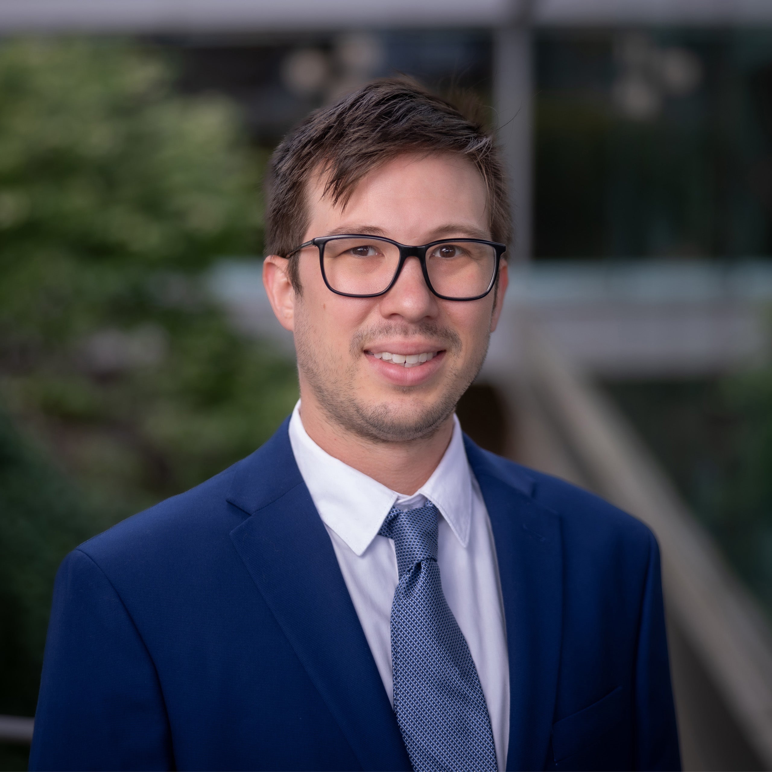 Jordan Taylor headshot. Male with short light brown hair, eyes, and wearing glasses. He is wearing a white shirt, blue tie, and blue suit jacket.
